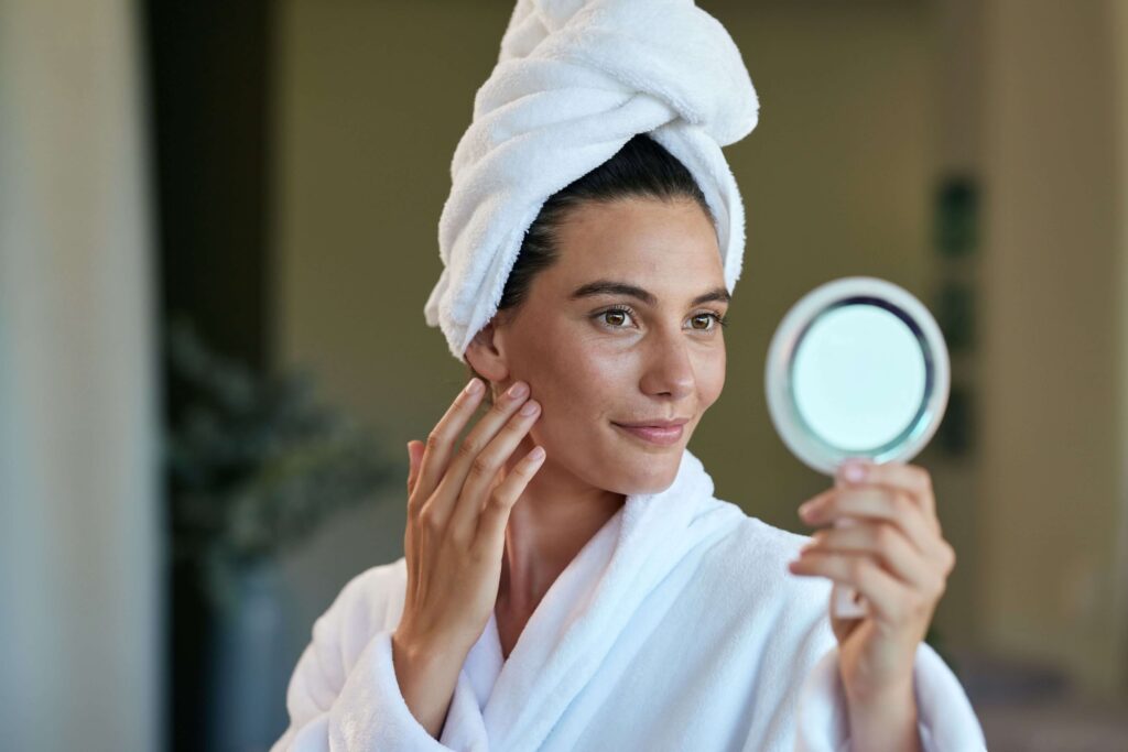 Woman in white robe examining her skin in a handheld mirror